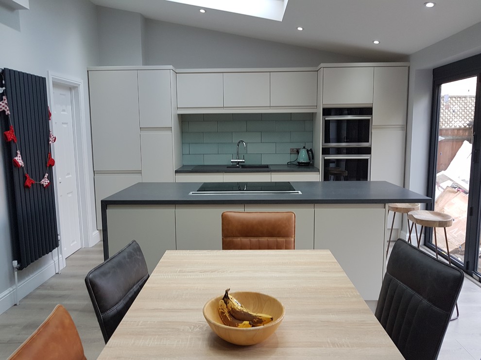 Photo of a mid-sized contemporary eat-in kitchen in Hertfordshire with an undermount sink, flat-panel cabinets, subway tile splashback, black appliances, laminate floors, with island and grey floor.