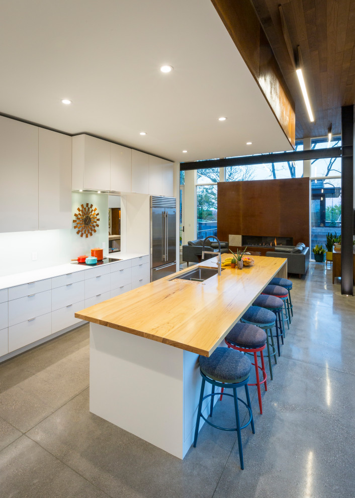Bild på ett funkis parallellkök, med en undermonterad diskho, släta luckor, vita skåp, träbänkskiva, rostfria vitvaror, betonggolv, en köksö, grått golv, vitt stänkskydd och glaspanel som stänkskydd
