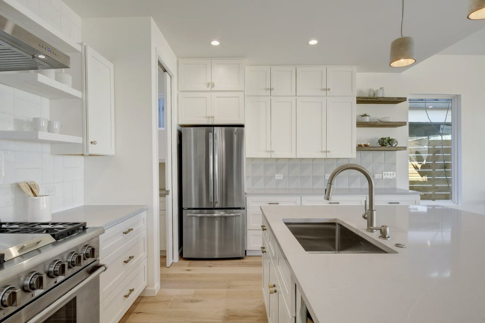 This is an example of a midcentury l-shaped open plan kitchen in Austin with an undermount sink, shaker cabinets, white cabinets, grey splashback, stainless steel appliances, medium hardwood floors and with island.