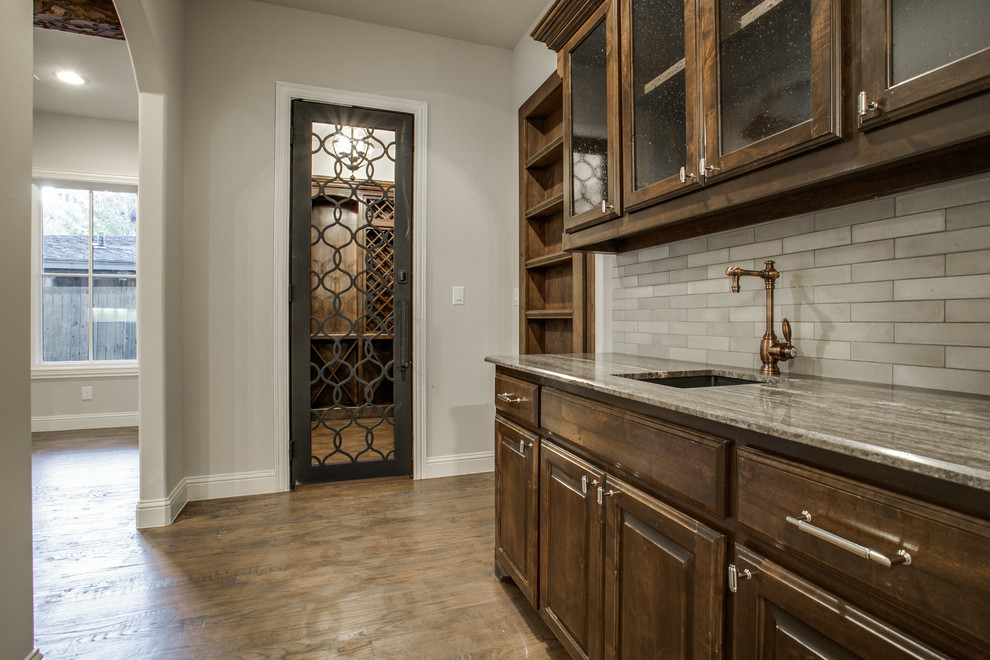 Photo of a large traditional galley wet bar in Dallas with an undermount sink, glass-front cabinets, dark wood cabinets, limestone benchtops, grey splashback, cement tile splashback and dark hardwood floors.