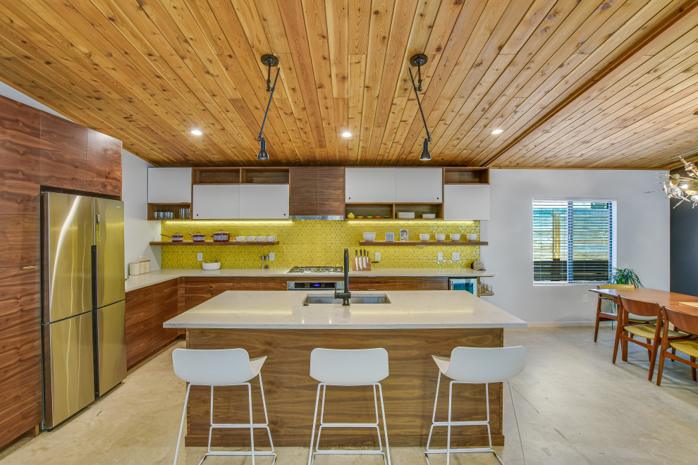 Bild på ett 60 tals vit vitt l-kök, med en undermonterad diskho, släta luckor, skåp i mellenmörkt trä, gult stänkskydd, rostfria vitvaror, betonggolv, en köksö och grått golv