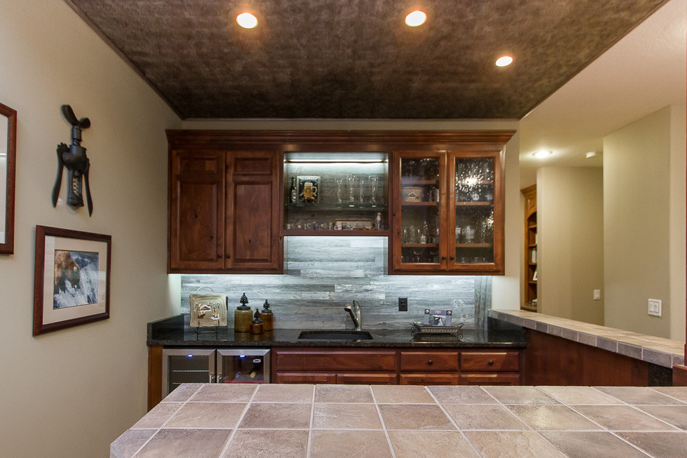 Inspiration for a mid-sized country u-shaped seated home bar in Kansas City with an undermount sink, raised-panel cabinets, dark wood cabinets, tile benchtops, grey splashback, timber splashback, medium hardwood floors and brown floor.