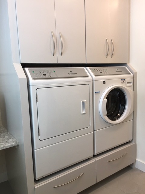 Photo of a mid-sized traditional single-wall dedicated laundry room in Portland Maine with flat-panel cabinets, grey cabinets, quartz benchtops, white walls, porcelain floors, a side-by-side washer and dryer, brown floor and a farmhouse sink.