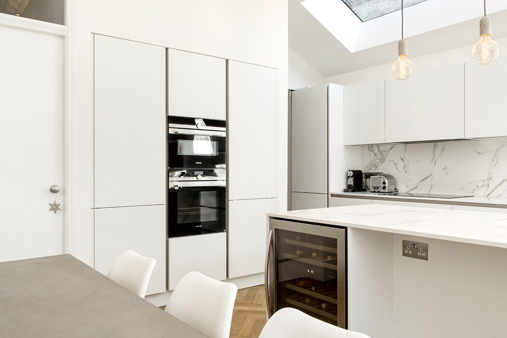 This is an example of a large contemporary l-shaped eat-in kitchen in London with flat-panel cabinets, white cabinets, white splashback, marble splashback, black appliances, light hardwood floors, with island, a single-bowl sink and quartzite benchtops.