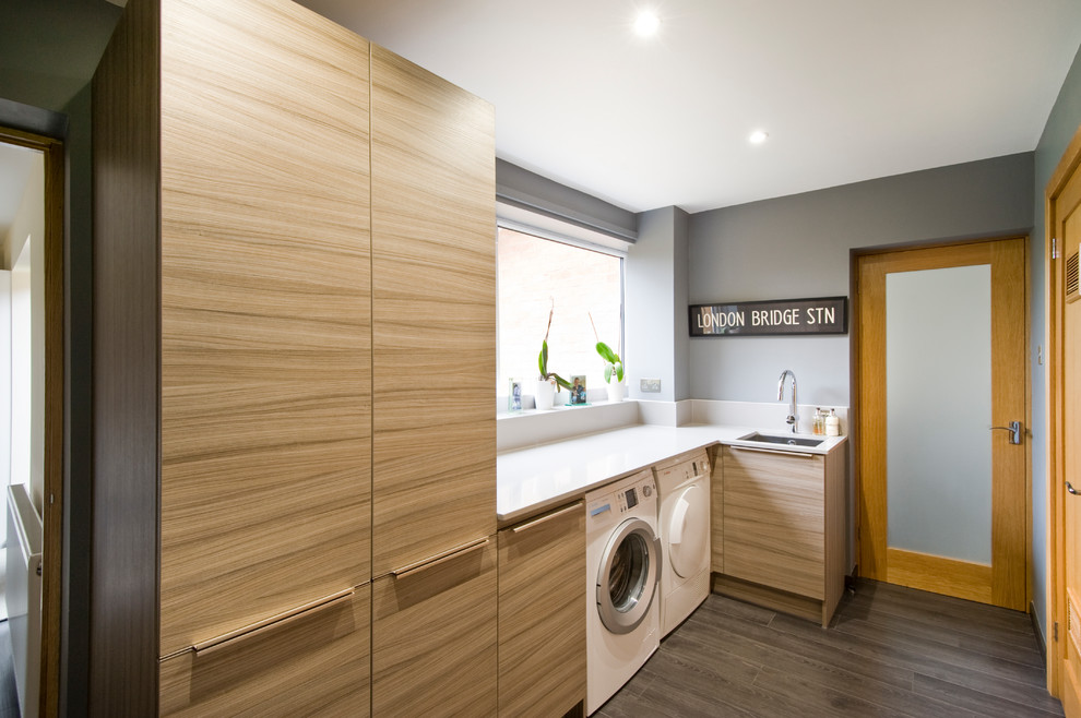 Mid-sized contemporary l-shaped utility room in West Midlands with a single-bowl sink, grey walls, laminate floors, a side-by-side washer and dryer, grey floor, white benchtop and light wood cabinets.