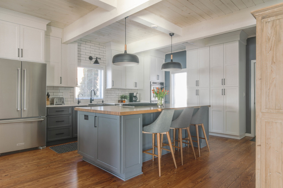 This is an example of a large country l-shaped eat-in kitchen with a farmhouse sink, recessed-panel cabinets, white cabinets, wood benchtops, white splashback, subway tile splashback, stainless steel appliances, medium hardwood floors, with island, brown floor and brown benchtop.