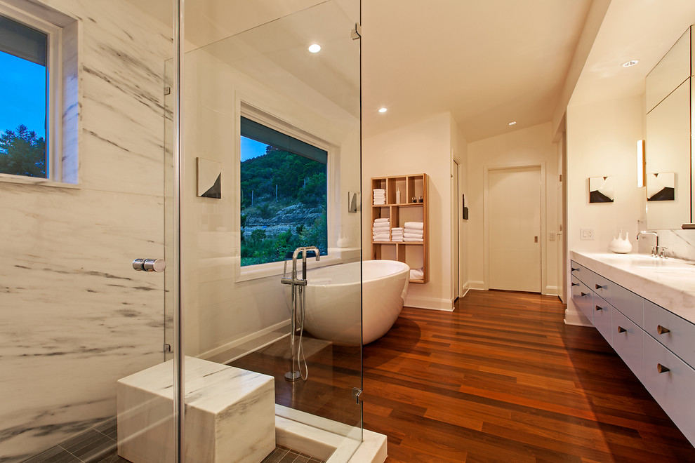 Design ideas for a large contemporary master bathroom in Austin with flat-panel cabinets, blue cabinets, a freestanding tub, an alcove shower, gray tile, white tile, marble, beige walls, dark hardwood floors, marble benchtops, brown floor and a hinged shower door.