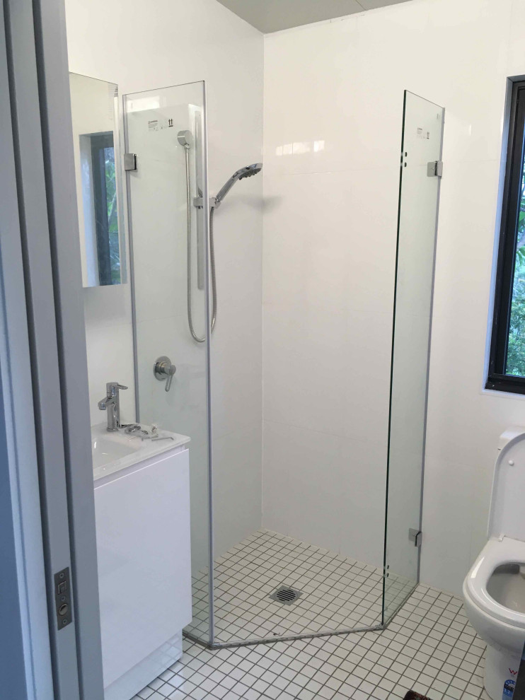 Small beach style master bathroom in Sunshine Coast with flat-panel cabinets, white cabinets, a corner shower, a one-piece toilet, white tile, ceramic tile, white walls, porcelain floors, an integrated sink, glass benchtops, white floor, a hinged shower door, white benchtops, a single vanity, a freestanding vanity and vaulted.
