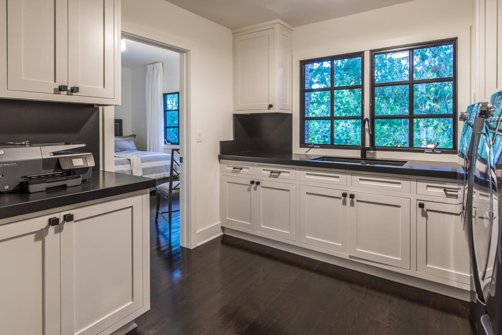This is an example of a traditional utility room in Los Angeles with a double-bowl sink, shaker cabinets, white cabinets, granite worktops, black splashback, granite splashback, white walls, dark hardwood flooring, a side by side washer and dryer, brown floors and black worktops.