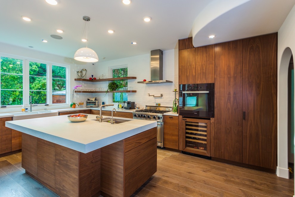 Photo of a mid-sized mediterranean l-shaped open plan kitchen in Los Angeles with a farmhouse sink, flat-panel cabinets, medium wood cabinets, solid surface benchtops, white splashback, brick splashback, stainless steel appliances, medium hardwood floors, with island and brown floor.
