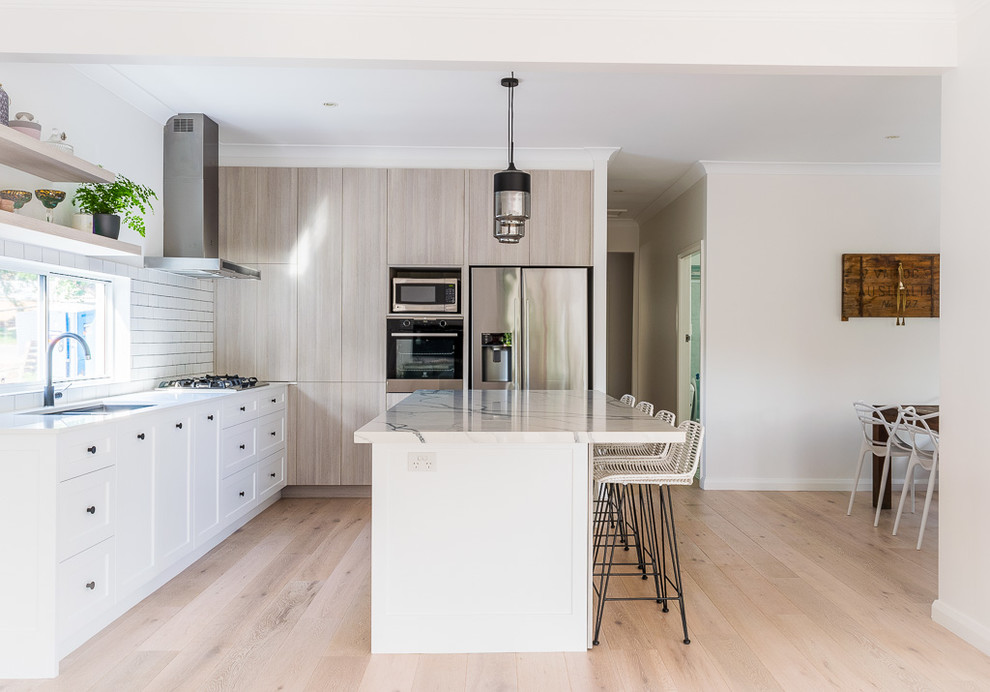 Mid-sized contemporary l-shaped open plan kitchen in Sydney with a double-bowl sink, quartz benchtops, white splashback, subway tile splashback, black appliances, light hardwood floors, with island, flat-panel cabinets, beige cabinets, beige floor and white benchtop.