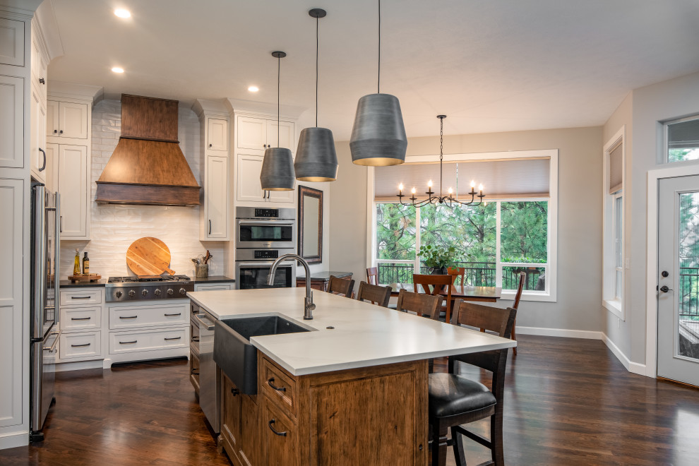Photo of a mid-sized transitional l-shaped eat-in kitchen in Seattle with a farmhouse sink, beaded inset cabinets, white cabinets, solid surface benchtops, white splashback, subway tile splashback, stainless steel appliances, medium hardwood floors, with island, brown floor and white benchtop.
