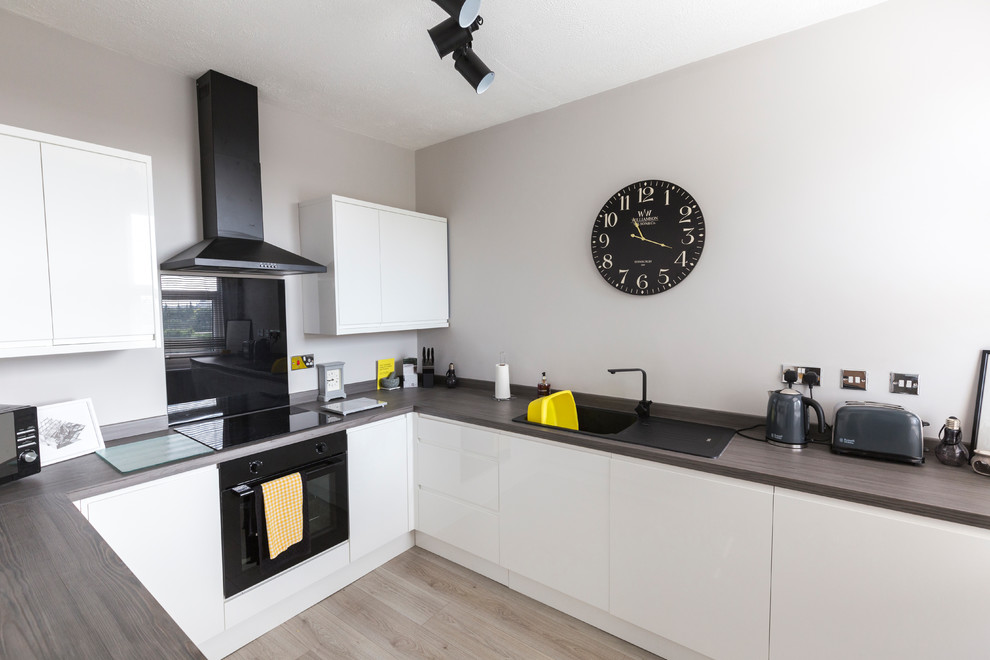 This is an example of a small contemporary u-shaped kitchen in Other with a drop-in sink, flat-panel cabinets, white cabinets, wood benchtops, black splashback, glass sheet splashback, black appliances, light hardwood floors, beige floor and brown benchtop.