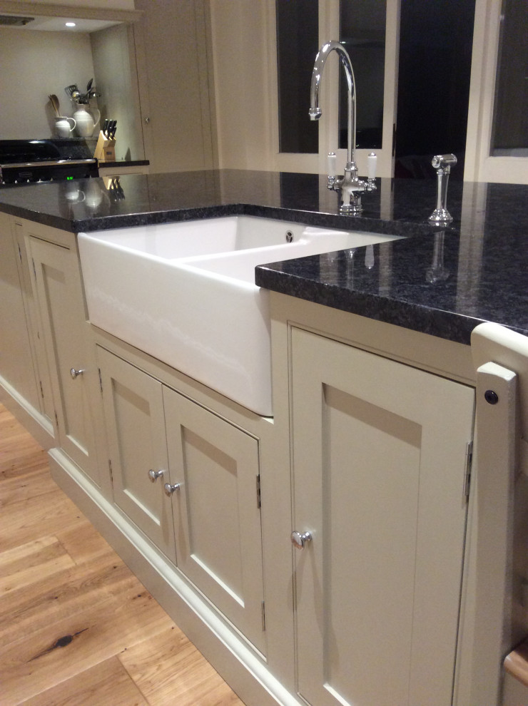 Photo of a small modern single-wall separate kitchen in Surrey with a farmhouse sink, beaded inset cabinets, beige cabinets, black appliances, light hardwood floors, with island and black benchtop.