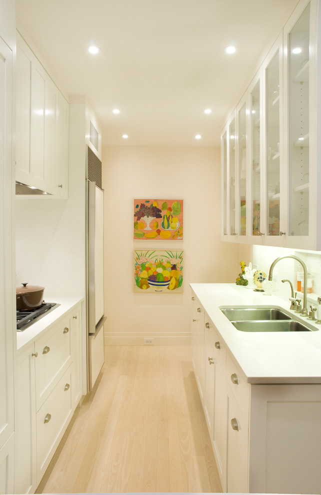 This is an example of a small transitional galley eat-in kitchen in New York with a double-bowl sink, glass-front cabinets, white cabinets, panelled appliances, solid surface benchtops, white splashback, ceramic splashback, light hardwood floors, no island and white floor.