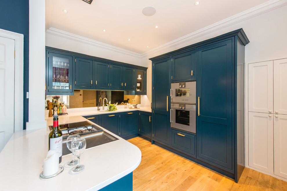 Inspiration for a mid-sized modern u-shaped eat-in kitchen in Edinburgh with a drop-in sink, shaker cabinets, blue cabinets, solid surface benchtops, mirror splashback, stainless steel appliances, light hardwood floors, a peninsula and white benchtop.