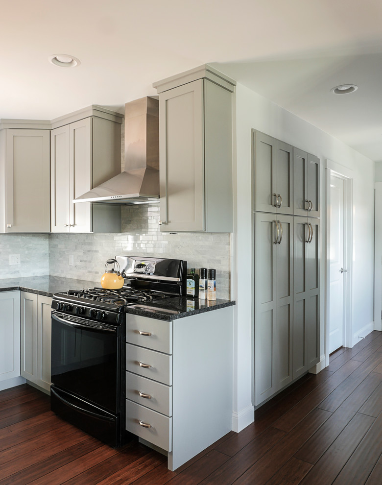 This is an example of a large transitional l-shaped eat-in kitchen in Burlington with a farmhouse sink, shaker cabinets, grey cabinets, granite benchtops, grey splashback, stone tile splashback, black appliances, bamboo floors and with island.