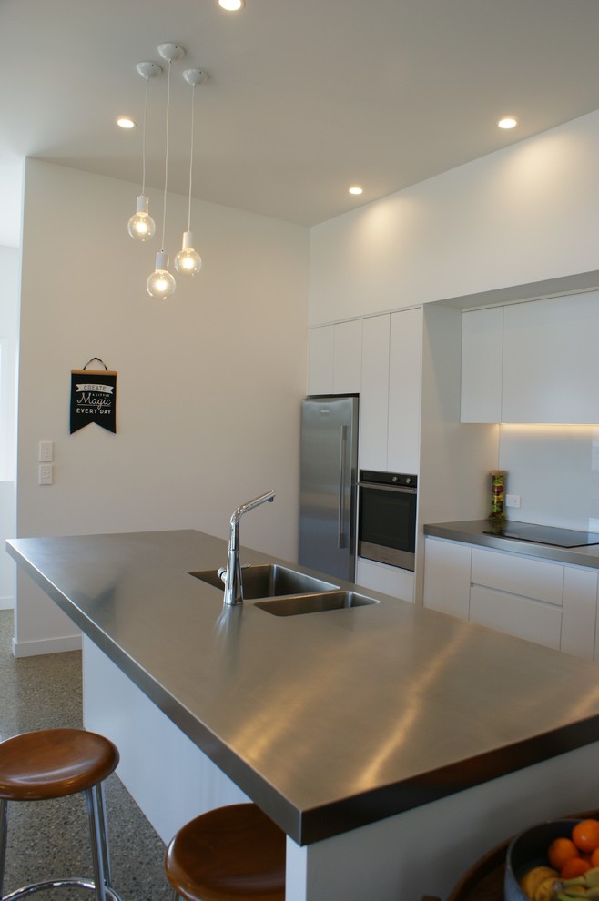 Design ideas for a large industrial galley kitchen pantry in Dunedin with an integrated sink, flat-panel cabinets, white cabinets, stainless steel benchtops, white splashback, glass sheet splashback, stainless steel appliances, concrete floors and with island.