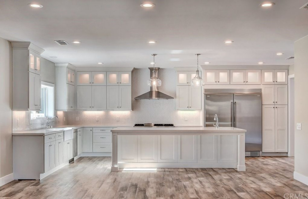 Large country l-shaped open plan kitchen in Orange County with a farmhouse sink, shaker cabinets, white cabinets, granite benchtops, white splashback, ceramic splashback, stainless steel appliances, light hardwood floors, with island, brown floor, multi-coloured benchtop and coffered.
