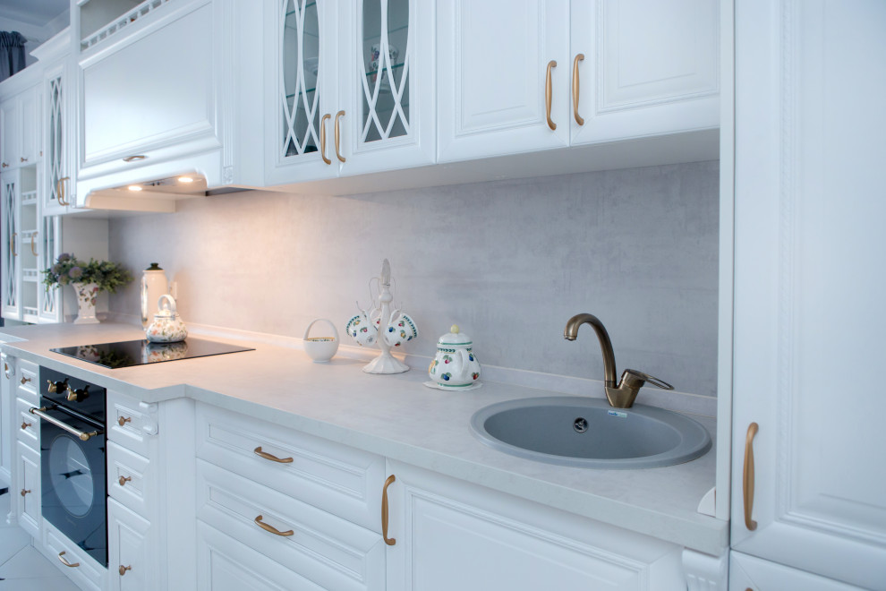Immagine di una cucina chic di medie dimensioni con lavello da incasso, ante con bugna sagomata, ante bianche, elettrodomestici neri, pavimento con piastrelle in ceramica, pavimento bianco e top grigio
