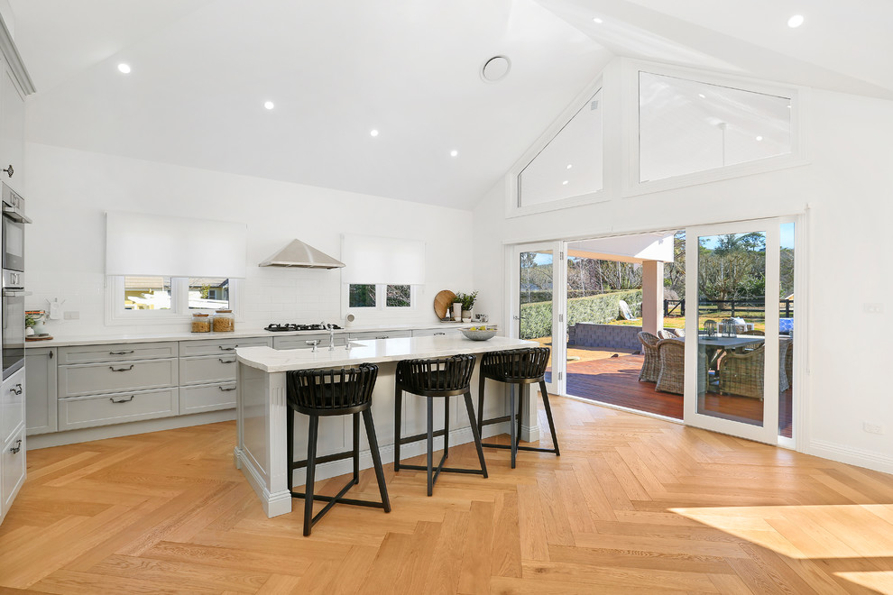 Design ideas for an expansive country l-shaped open plan kitchen in Wollongong with a farmhouse sink, shaker cabinets, grey cabinets, quartz benchtops, white splashback, ceramic splashback, stainless steel appliances, with island, medium hardwood floors, brown floor and white benchtop.