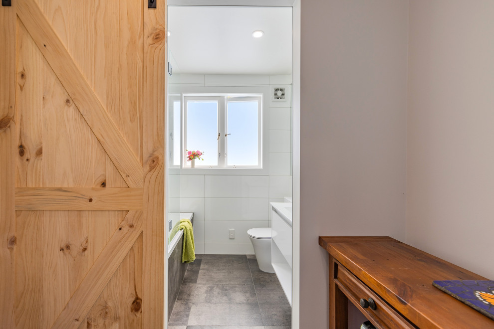 Photo of a small modern 3/4 bathroom in Auckland with flat-panel cabinets, white cabinets, a drop-in tub, a shower/bathtub combo, a one-piece toilet, white tile, ceramic tile, white walls, porcelain floors, an integrated sink, solid surface benchtops, grey floor, a hinged shower door, white benchtops, a single vanity and a built-in vanity.