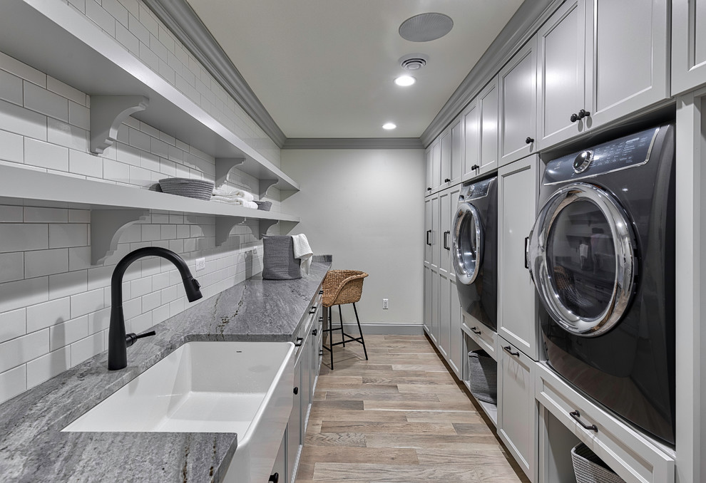 Design ideas for a large transitional galley dedicated laundry room in Detroit with a farmhouse sink, recessed-panel cabinets, grey cabinets, white walls, light hardwood floors, an integrated washer and dryer, beige floor, grey benchtop and granite benchtops.
