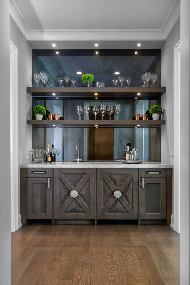 This is an example of a large transitional wet bar in Detroit with distressed cabinets, marble benchtops, mirror splashback, medium hardwood floors and brown floor.