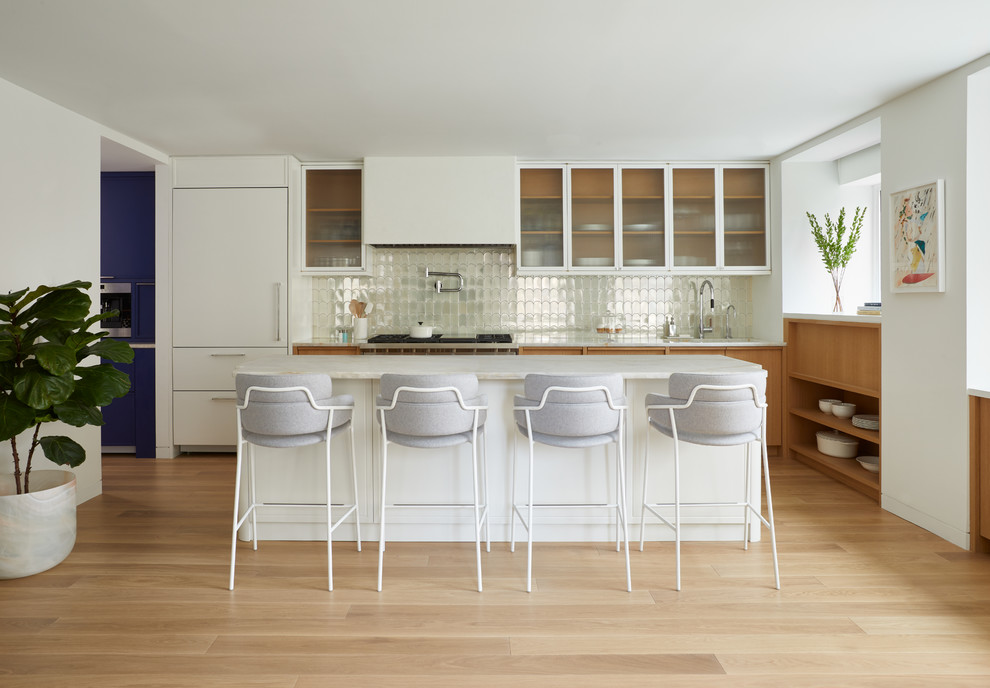 This is an example of a contemporary open plan kitchen in New York with glass-front cabinets, white cabinets, metallic splashback, metal splashback, panelled appliances, light hardwood floors, with island, beige floor and white benchtop.