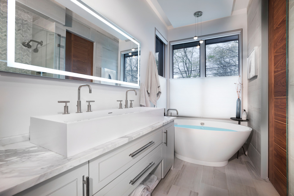 This is an example of a mid-sized contemporary master bathroom in Other with raised-panel cabinets, white cabinets, a freestanding tub, a corner shower, a one-piece toilet, gray tile, marble, grey walls, light hardwood floors, a trough sink, marble benchtops, beige floor, a hinged shower door and grey benchtops.