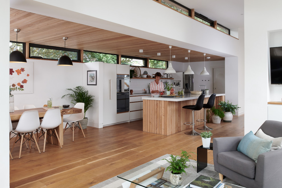 Inspiration for a mid-sized contemporary single-wall open plan kitchen in Cambridgeshire with flat-panel cabinets, white cabinets, recycled glass benchtops, white splashback, ceramic splashback, stainless steel appliances, medium hardwood floors, with island and multi-coloured benchtop.