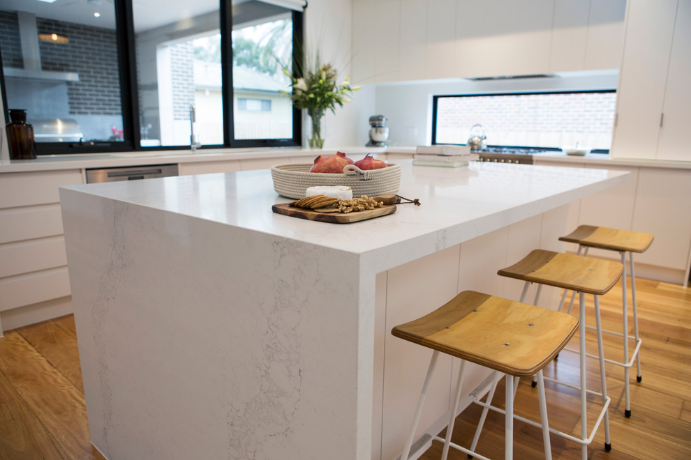 This is an example of a large contemporary l-shaped eat-in kitchen in Melbourne with a double-bowl sink, flat-panel cabinets, white cabinets, quartz benchtops, window splashback, white appliances, medium hardwood floors and with island.