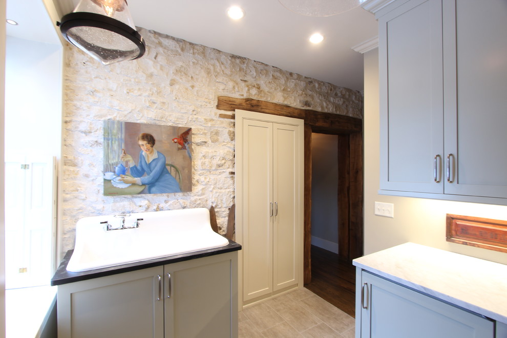 Photo of a mid-sized country galley utility room in Other with a farmhouse sink, recessed-panel cabinets, granite benchtops, beige walls, porcelain floors, a stacked washer and dryer, brown floor, green benchtop and grey cabinets.