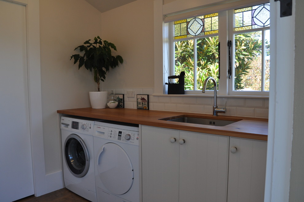 This is an example of a mid-sized transitional single-wall dedicated laundry room in Auckland with an undermount sink, white cabinets, wood benchtops, beige walls, dark hardwood floors and a side-by-side washer and dryer.