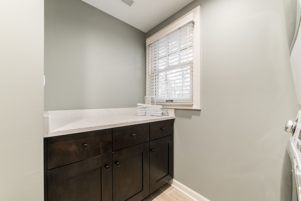 This is an example of a large traditional galley dedicated laundry room in Chicago with brown cabinets, quartzite benchtops, grey walls, ceramic floors, a stacked washer and dryer, beige floor and white benchtop.