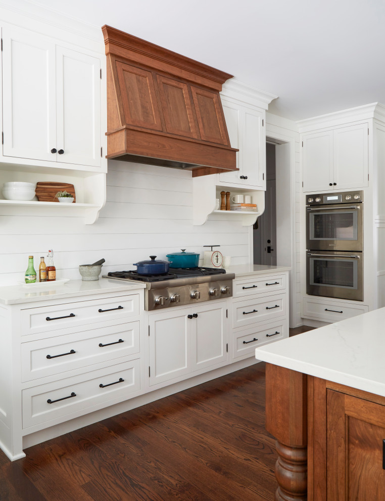 This is an example of a large rural u-shaped open plan kitchen in Chicago with a belfast sink, recessed-panel cabinets, white cabinets, engineered stone countertops, white splashback, tonge and groove splashback, stainless steel appliances, medium hardwood flooring, an island, brown floors and white worktops.