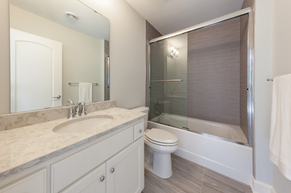 Photo of a mid-sized traditional 3/4 bathroom in Minneapolis with recessed-panel cabinets, white cabinets, an alcove tub, a shower/bathtub combo, a two-piece toilet, gray tile, ceramic tile, white walls, ceramic floors, an undermount sink, engineered quartz benchtops, grey floor and a sliding shower screen.