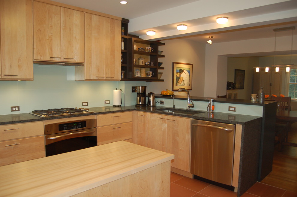 This is an example of a mid-sized contemporary l-shaped eat-in kitchen in DC Metro with an undermount sink, flat-panel cabinets, light wood cabinets, granite benchtops, white splashback, glass tile splashback, stainless steel appliances, terra-cotta floors, a peninsula, red floor and black benchtop.