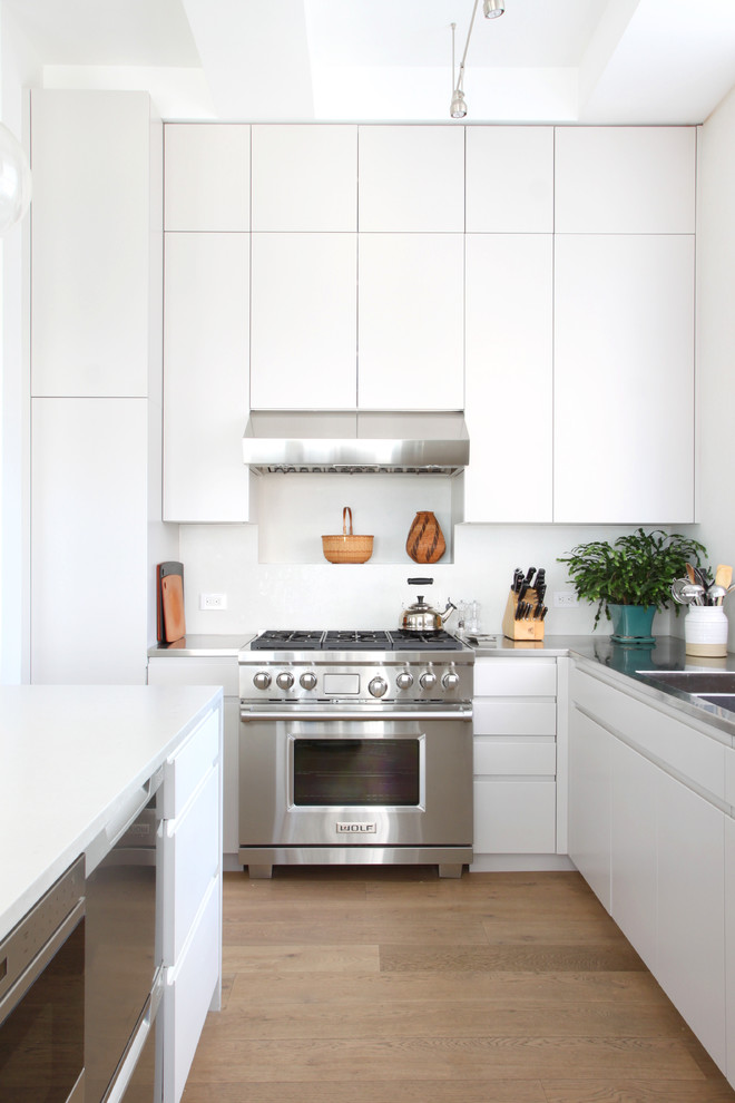 Inspiration for a small contemporary l-shaped kitchen in New York with a double-bowl sink, flat-panel cabinets, white splashback, light hardwood floors, with island, white cabinets, stainless steel benchtops and stainless steel appliances.