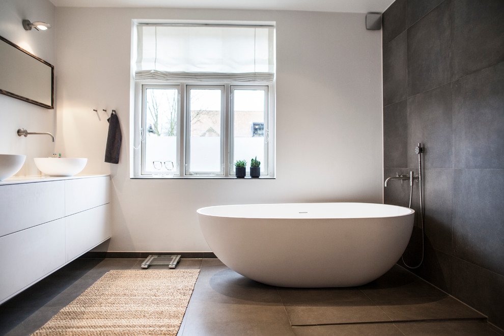 Photo of a large modern master bathroom in Wiltshire with flat-panel cabinets, white cabinets, a freestanding tub, a shower/bathtub combo, gray tile, stone tile, white walls and a vessel sink.