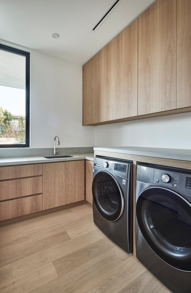 Inspiration for a large modern l-shaped dedicated laundry room in Los Angeles with an undermount sink, brown cabinets, solid surface benchtops, grey splashback, engineered quartz splashback, white walls, light hardwood floors, a side-by-side washer and dryer, yellow floor and grey benchtop.