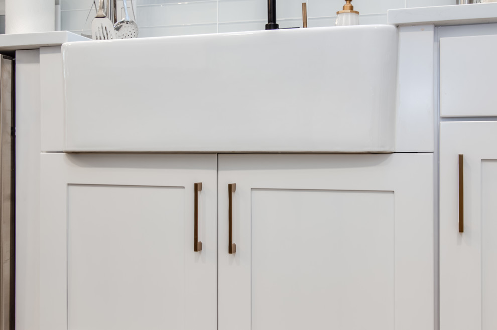 Example of a large transitional l-shaped porcelain tile and gray floor kitchen pantry design in Houston with a farmhouse sink, shaker cabinets, white cabinets, quartzite countertops, white backsplash, glass tile backsplash, stainless steel appliances, an island and white countertops