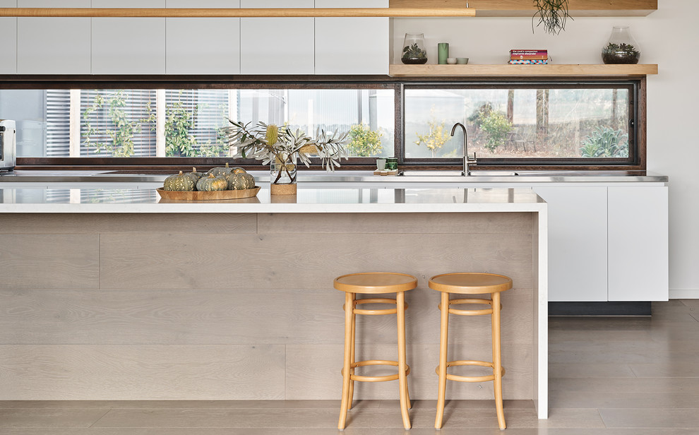 Photo of a large contemporary galley kitchen in Melbourne with flat-panel cabinets, white cabinets, window splashback, medium hardwood floors, with island, brown floor and white benchtop.
