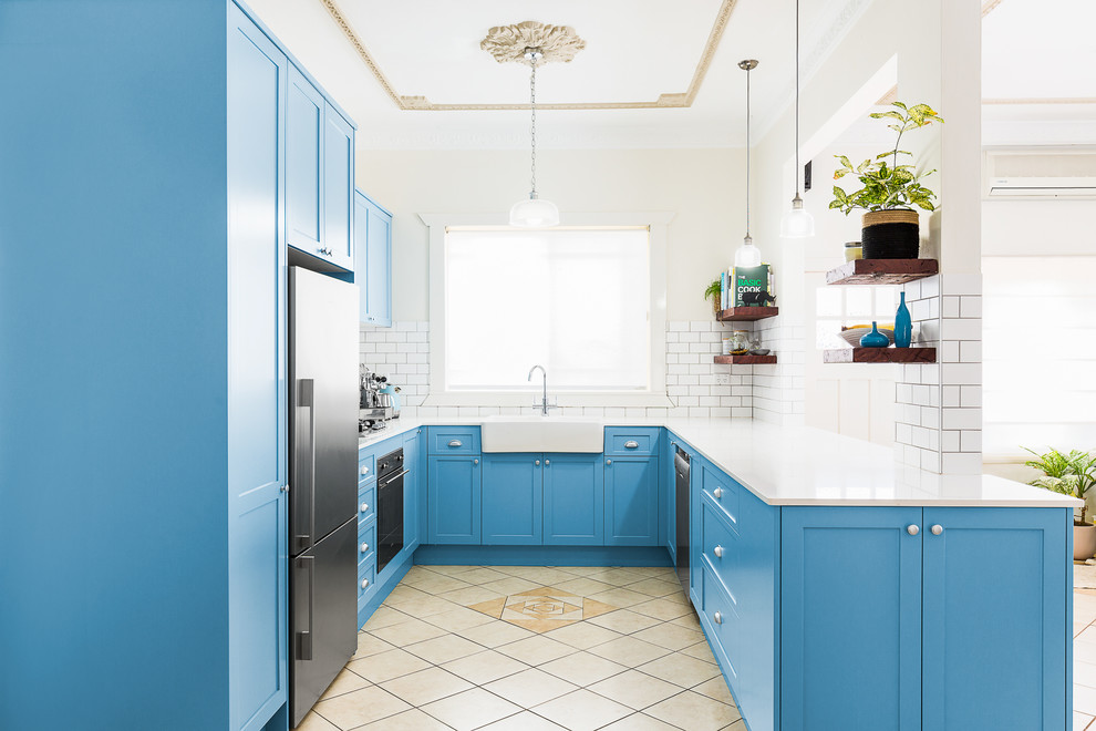 Inspiration for a mid-sized transitional u-shaped kitchen in Sydney with a farmhouse sink, blue cabinets, white splashback, subway tile splashback, stainless steel appliances, beige floor, shaker cabinets, solid surface benchtops, terra-cotta floors, with island and white benchtop.