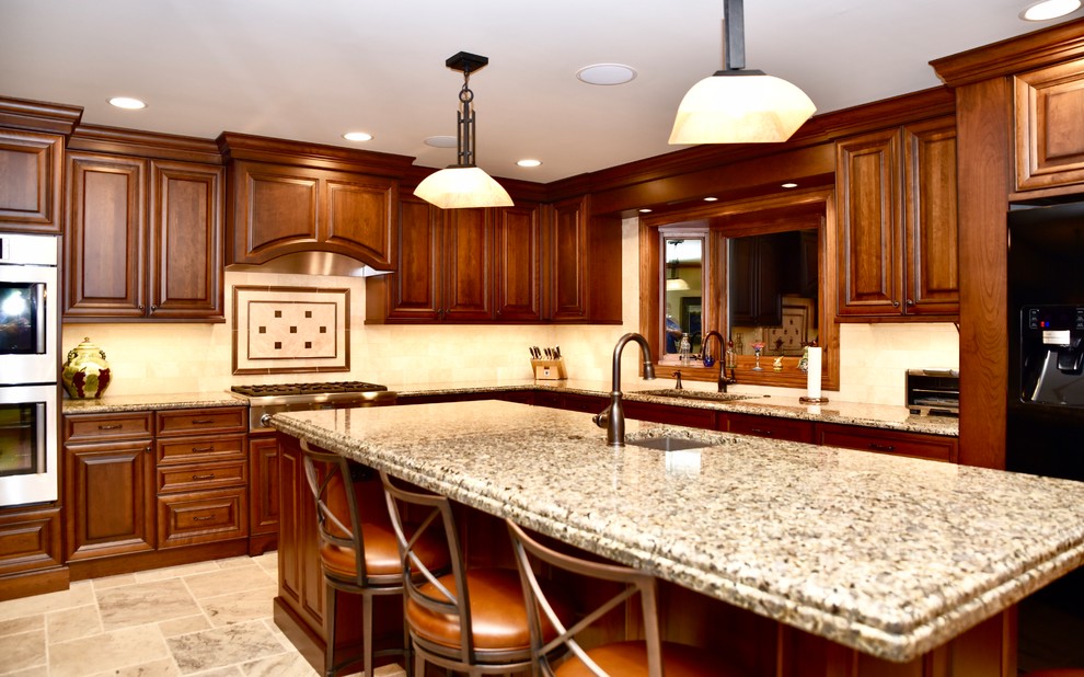 This is an example of a large traditional u-shaped eat-in kitchen in Chicago with a double-bowl sink, raised-panel cabinets, medium wood cabinets, granite benchtops, beige splashback, travertine splashback, black appliances, travertine floors, with island, beige floor and beige benchtop.
