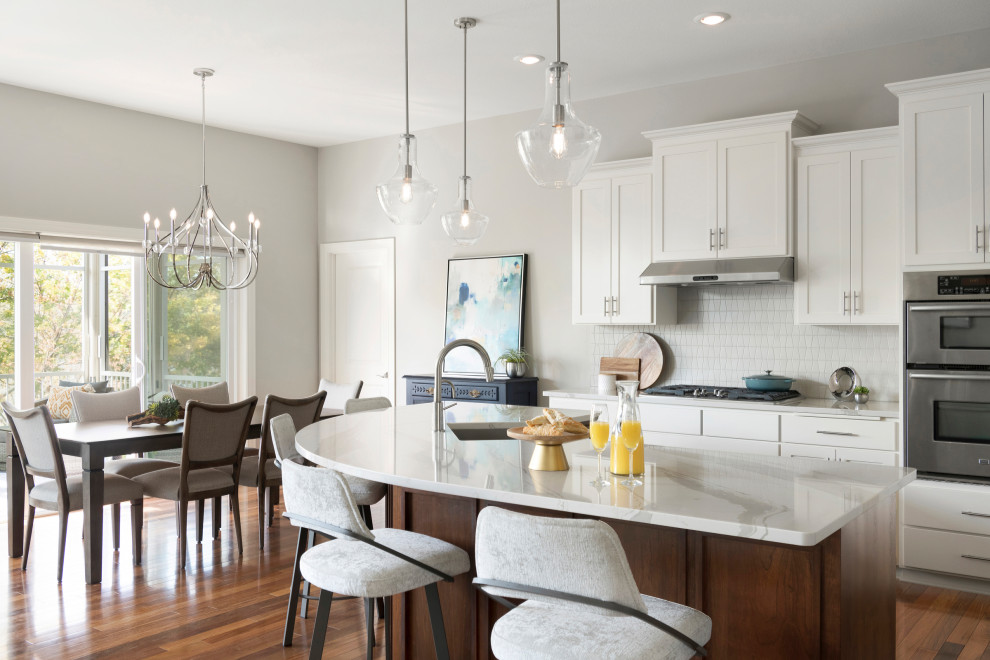 Large beach style u-shaped eat-in kitchen in Minneapolis with a drop-in sink, recessed-panel cabinets, white cabinets, quartzite benchtops, white splashback, ceramic splashback, stainless steel appliances, medium hardwood floors, with island, brown floor and white benchtop.