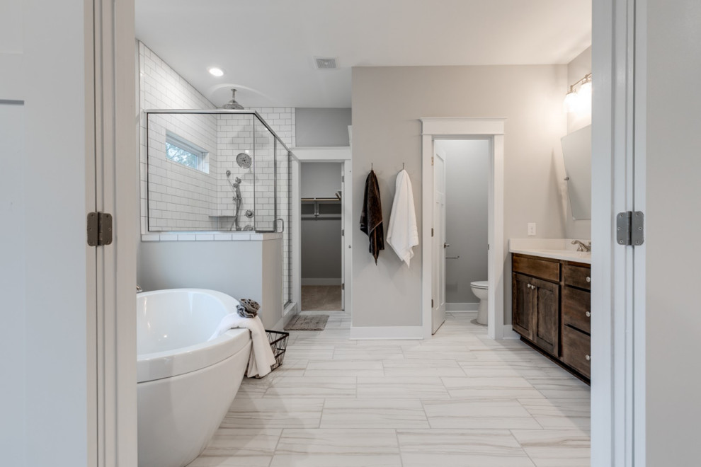 Photo of a large arts and crafts master bathroom in Richmond with recessed-panel cabinets, brown cabinets, a freestanding tub, a corner shower, a two-piece toilet, white tile, subway tile, grey walls, ceramic floors, an undermount sink, solid surface benchtops, white floor, a hinged shower door, white benchtops, an enclosed toilet, a double vanity and a built-in vanity.