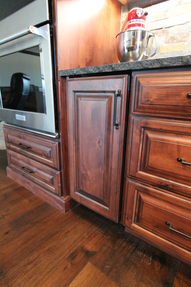 Large mountain style medium tone wood floor and brown floor kitchen photo with raised-panel cabinets, medium tone wood cabinets, granite countertops, multicolored backsplash, stone tile backsplash, paneled appliances, an island and black countertops