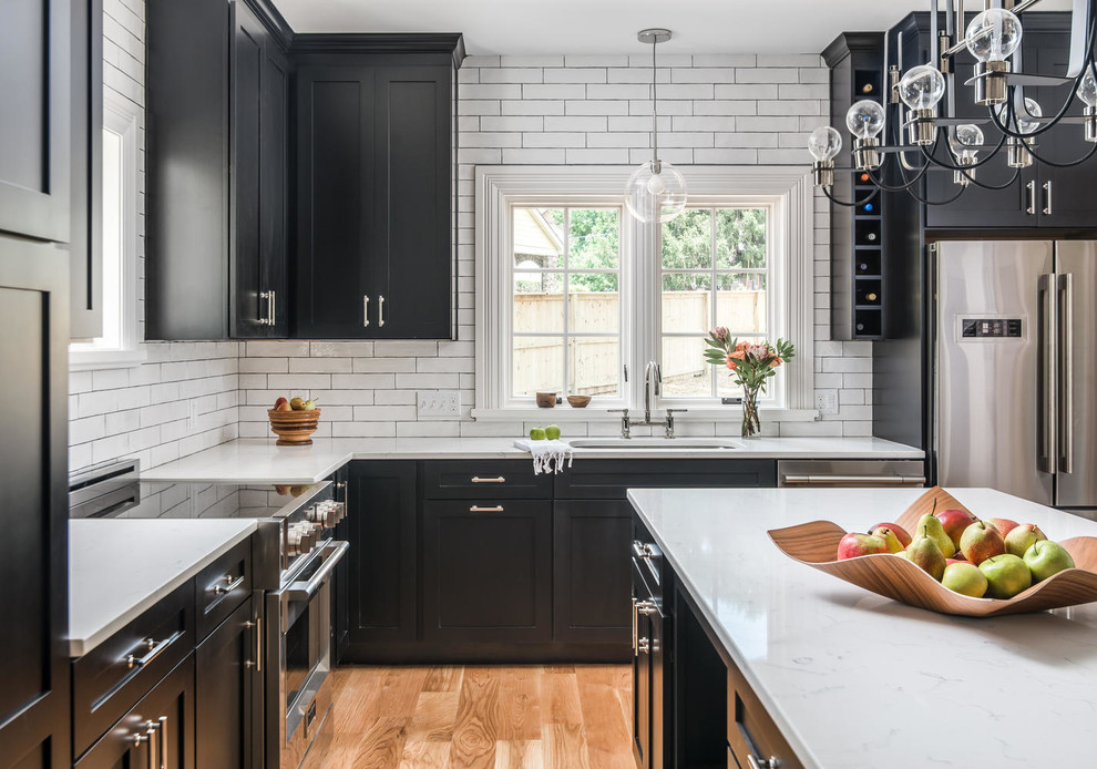 This is an example of a large transitional l-shaped open plan kitchen in Nashville with an undermount sink, shaker cabinets, black cabinets, quartzite benchtops, white splashback, ceramic splashback, stainless steel appliances, light hardwood floors, with island and white benchtop.