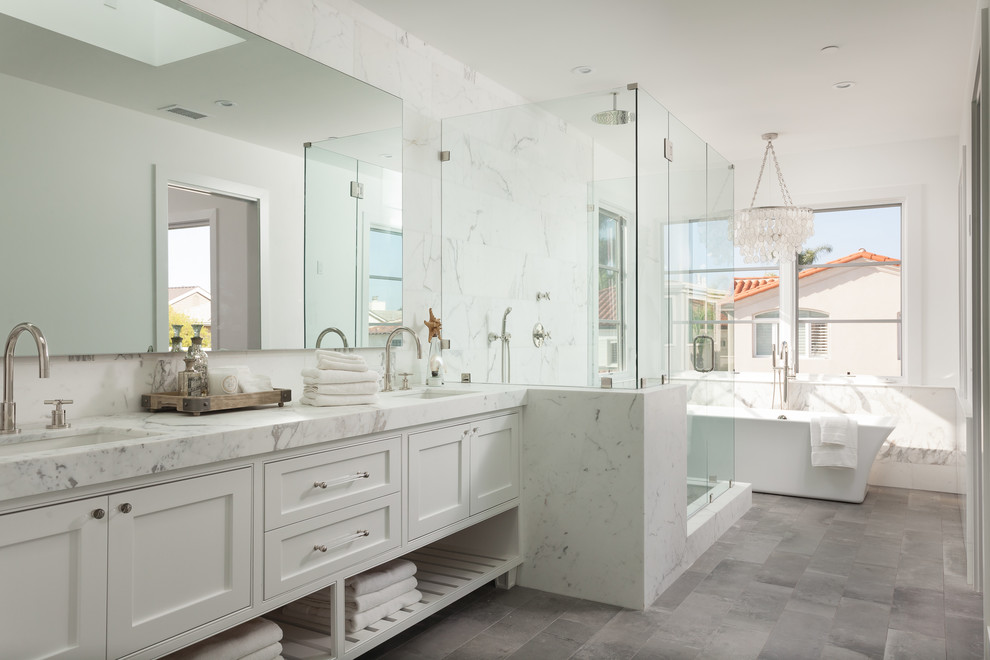 This is an example of a large beach style master bathroom in Orange County with an undermount sink, white cabinets, marble benchtops, a freestanding tub, white tile, stone tile, white walls, limestone floors, shaker cabinets, a corner shower and grey floor.
